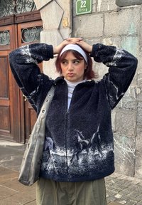 Veste en polaire bleu marine foncé, présentant une finition texturée et une impression graphique de chevaux le long de l'ourlet. Accessoirisée d'un sac à bandoulière gris métallique.