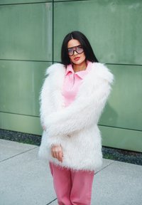 Bílý kabát z umělé kožešiny, růžový pletený top s límcem, růžové širší kalhoty, přehnaně velké černé sluneční brýle, stojící proti zelené zdi.