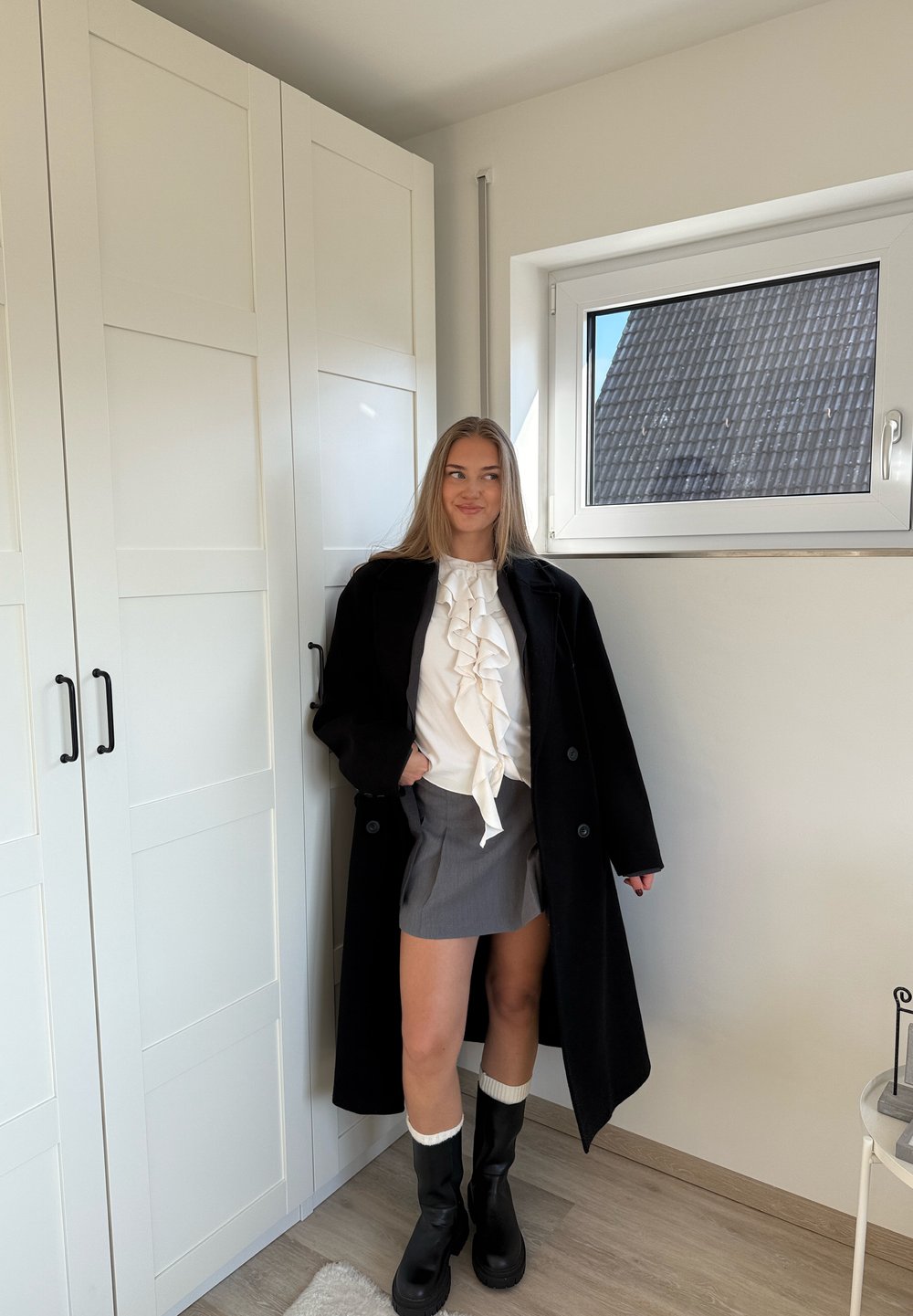 Abrigo negro, blusa blanca con volantes, falda gris, botas negras a la altura de la rodilla con puños blancos. Ambientación moderna con paredes claras y armarios.