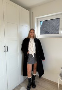 Abrigo negro, blusa blanca con volantes, falda gris, botas negras a la altura de la rodilla con puños blancos. Ambientación moderna con paredes claras y armarios.