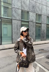 Olivově zelená kožená bunda, bílé tričko, krémové kalhoty, béžová čepice s logem NY, nadměrné černé sluneční brýle a tmavá kabelka s akcenty.