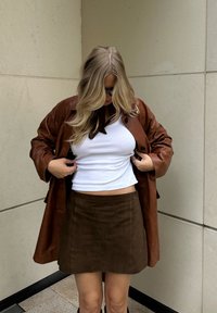 Bruine leren jas, witte tanktop en een bruin suède minirok. Lang haar, minimale sieraden en een eenvoudige sjaal maken de outfit compleet.