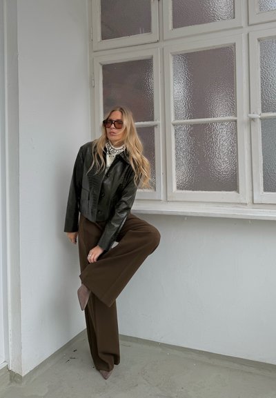 Mujer con cabello largo y rubio, usando gafas de sol, una chaqueta negra, pantalones marrones de pierna ancha y zapatos marrones puntiagudos, de pie en el interior junto a ventanas texturizadas.