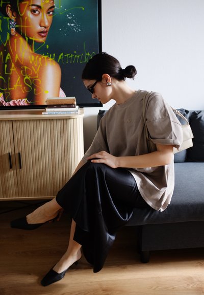 Camiseta beige de gran tamaño, falda de satén negra y zuecos negros. Sentada en un sofá oscuro con un armario de madera y libros de fondo.