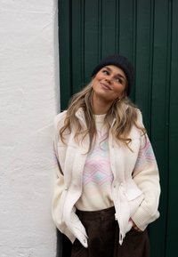 Jeune femme en gilet polaire blanc, pull à motifs, bonnet noir et pantalon foncé, appuyée contre un mur, souriant et regardant en hauteur sur le côté.