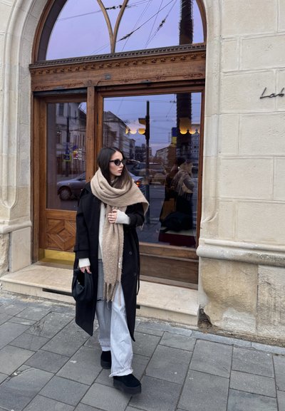 Mujer con abrigo negro, bufanda beige, gafas de sol y jeans claros de pie en la acera de la ciudad frente a una puerta de vidrio con marco de madera.