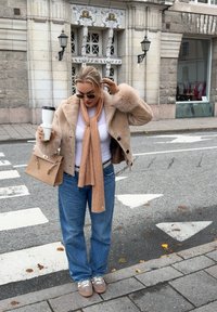 Veste beige bordée de fourrure, chemise blanche, écharpe tricotée marron clair, jean bleu ample, sac marron à motifs, baskets marron ; cadre urbain.