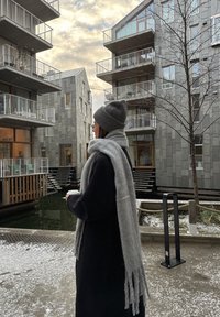 Cachecol e gorro de lã cinza usados por uma pessoa com um casaco preto, de pé perto de edifícios modernos com varandas de madeira, rodeada de neve.