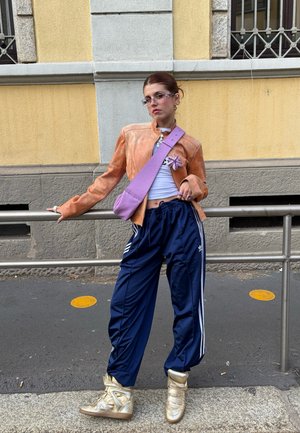 Chaqueta de cuero marrón claro, top corto blanco, pantalones deportivos navy con rayas blancas, zapatillas altas doradas y bolso cruzado morado.