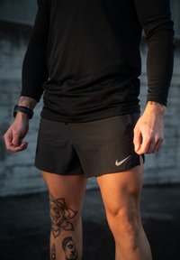 Αθλητής που φοράει μαύρο μακρυμάνικο μπλουζάκι και μαύρα σορτς Nike, με τατουάζ στο αριστερό πόδι και ρολόι στον αριστερό καρπό, στέκεται σε εξωτερικό χώρο.