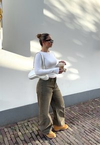 Hvit langermet topp, olivengrønne vide bukser, gule støvler og hvit veske. Holder en drink, står mot en lys vegg.