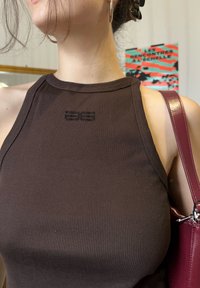 Femme portant un top sans manches côtelé marron avec un petit logo noir, portant un sac à bandoulière bordeaux, cheveux attachés en arrière et boucles d'oreilles créoles dorées.
