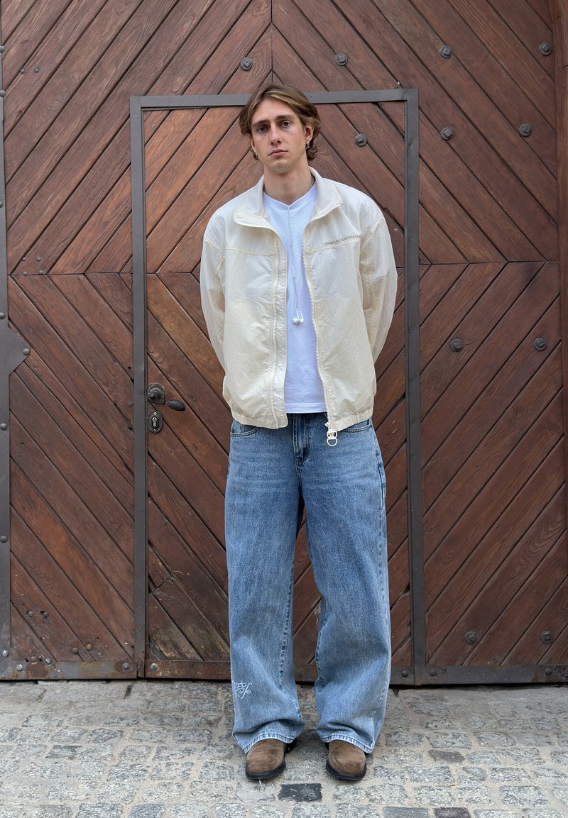 Jeune homme les mains derrière le dos, portant une veste blanche, une chemise blanche, un jean bleu ample et des chaussures marron, debout devant une grande porte en bois.