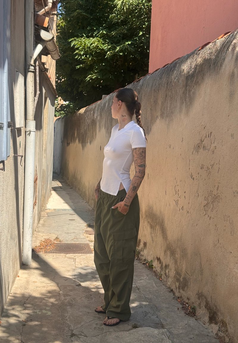 Personne avec des cheveux tressés et des tatouages se tient dans une ruelle étroite ensoleillée, portant une chemise blanche, un pantalon cargo vert et des sandales noires.