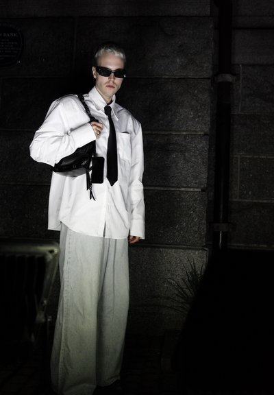 Camisa blanca de gran tamaño, corbata negra y jeans de pierna ancha color azul claro. Los accesorios incluyen un bolso negro de hombro y gafas de sol oscuras. Fondo urbano.