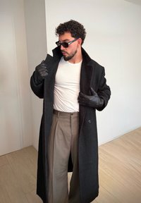 Manteau noir à larges revers, porté par-dessus un t-shirt blanc. Pantalon gris plissé, gants noirs et lunettes de soleil complètent la tenue.