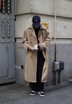 Beige Trenchcoat mit großen Knöpfen, getragen über einem schwarzen Hemd und dunklen Hosen. Schwarze Sneakers mit weißen Sohlen. Hält eine Kaffeetasse.