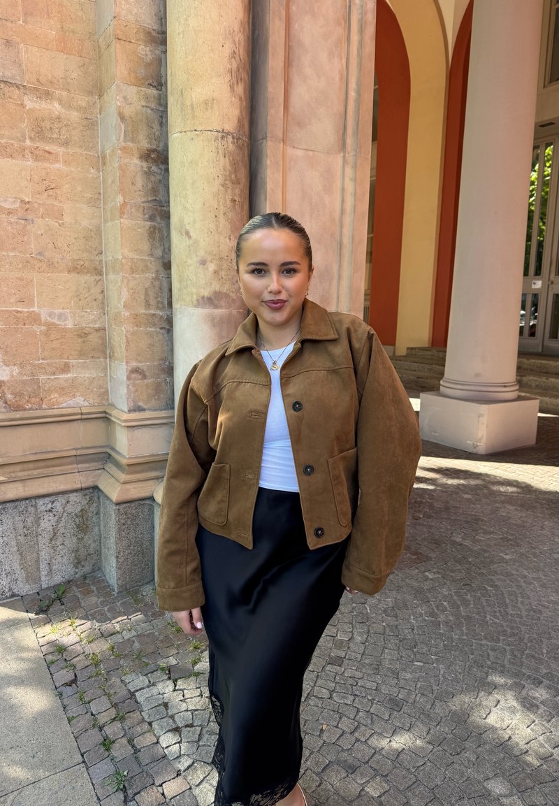 Jeune femme debout près de grandes colonnes en pierre, portant une veste marron, un haut blanc, une jupe noire et un collier doré dans un cadre extérieur ensoleillé.