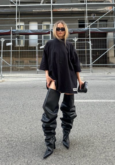 Camiseta negra de gran tamaño, botas de cuero negras hasta la rodilla con un diseño desgarbado y un bolso negro. Fondo urbano con andamios de construcción.