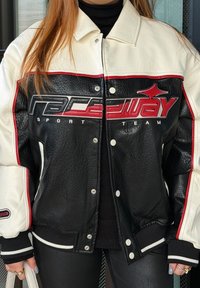 Personne portant une veste varsity en cuir noir et blanc avec un logo rouge « Raceway Sport Team », un col roulé noir et un pantalon noir, tenant un sac blanc.