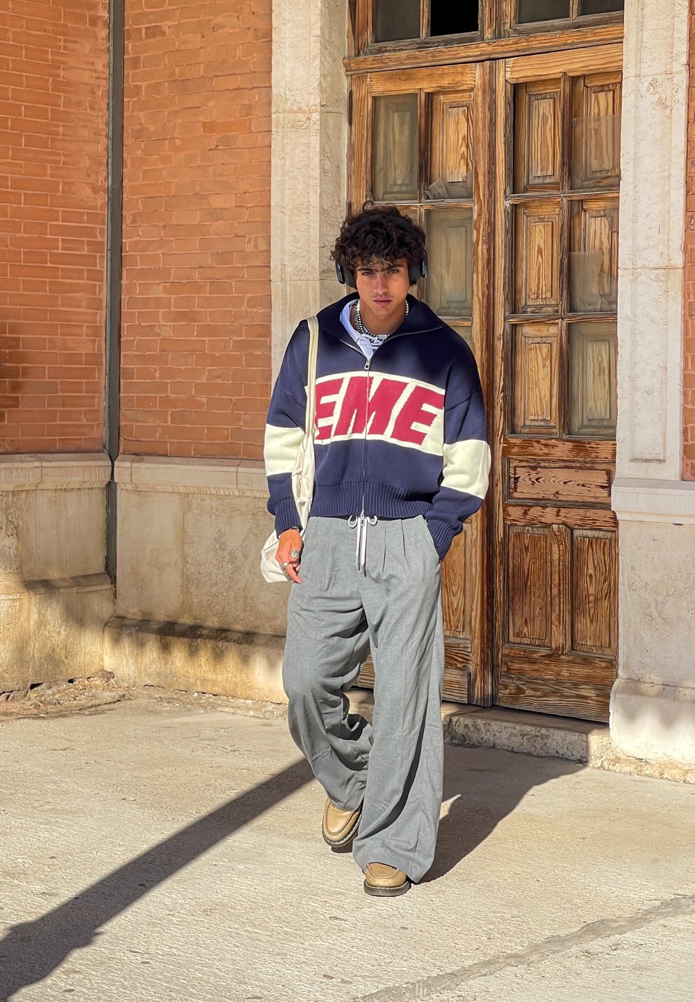 Ναυτικό και κρεμ πουλόβερ με το "EME" σε κόκκινα γράμματα, γκρι παντελόνια wide-leg, καφέ παπούτσια και ακουστικά, στέκεται κοντά σε ξύλινες πόρτες.
