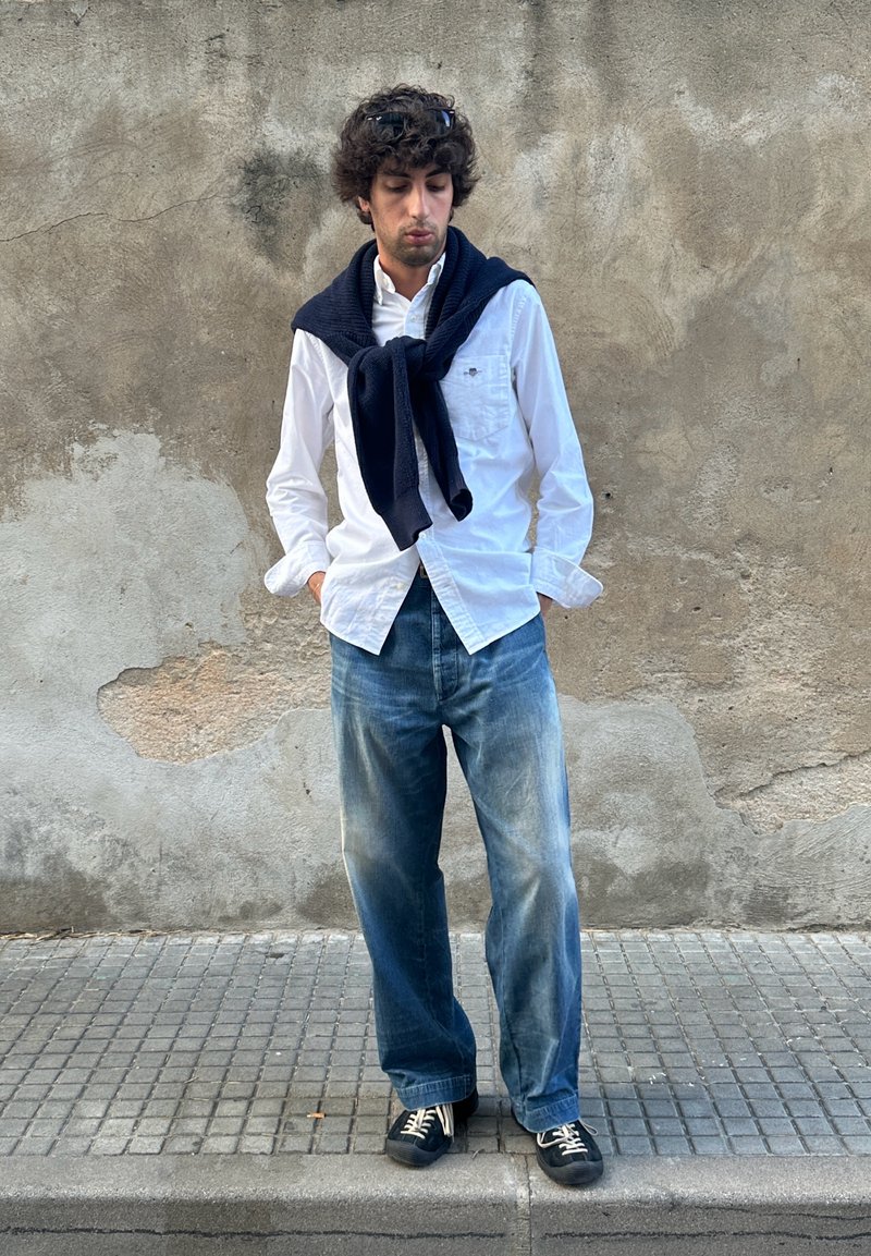 Junger Mann steht auf dem Gehsteig, trägt ein weißes Hemd, blaue Jeans, schwarze Sneakers, Sonnenbrille auf lockigem Haar und einen marineblauen Pullover über den Schultern.