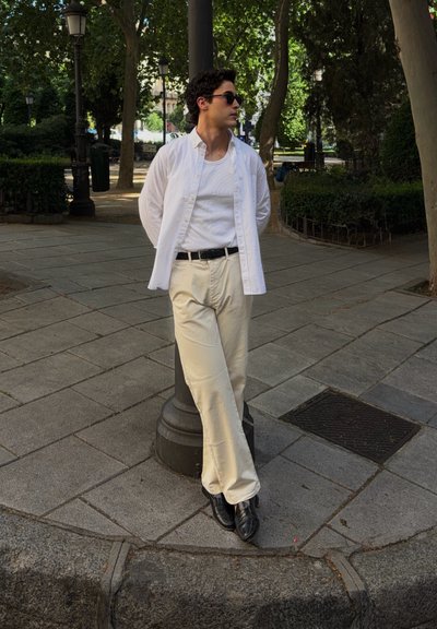 Joven con gafas de sol, camisa blanca y pantalones beige apoyado en un farol en un parque urbano bordeado de árboles.