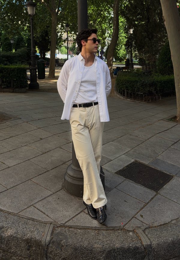 Jeune homme portant des lunettes de soleil, une chemise blanche et un pantalon beige appuyé contre un lampadaire dans un parc urbain bordé d'arbres.
