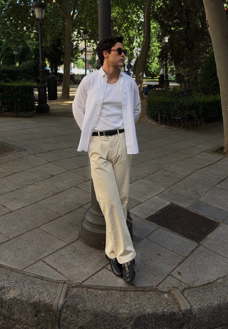 Jeune homme portant des lunettes de soleil, une chemise blanche et un pantalon beige appuyé contre un réverbère dans un parc urbain bordé d'arbres.