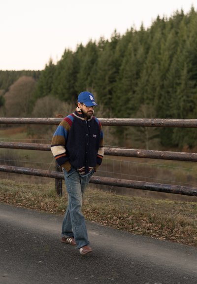 Cardigan de punto con rayas multicolores, base azul marino y botones grandes. Llevado sobre una camisa azul, combinado con jeans claros y zapatos marrones.