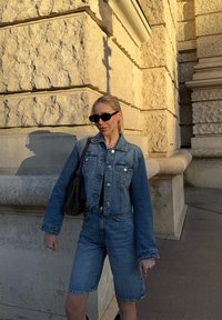 Traper jakna s krojenjem do struka, plave boje, s prednjim gumbima, uparen s odgovarajućim traper šortsevima. Crni dodaci kontrastiraju sa odjevnom kombinacijom.