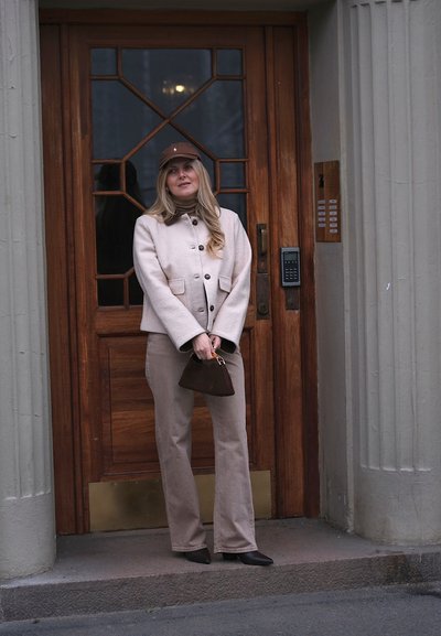 Mujer con chaqueta y pantalones beige, gorra marrón, sosteniendo un bolso oscuro, de pie frente a una puerta de madera con paneles de vidrio y teclado de seguridad.