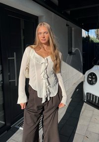 Blusa bianca con volant, maniche lunghe e scollo a V, abbinata a pantaloni marrone scuro larghi. Pieghe sottili sul tessuto della blusa.