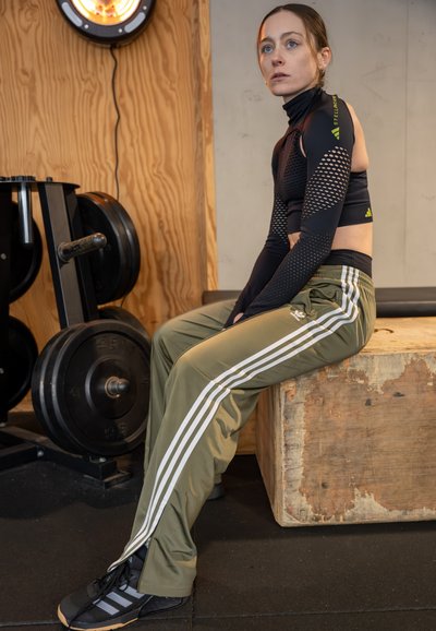 Top corto de manga larga negro con detalles de malla, pantalones deportivos verde oliva con franjas blancas y zapatillas de entrenamiento negras. Banco de madera en un entorno de gimnasio.