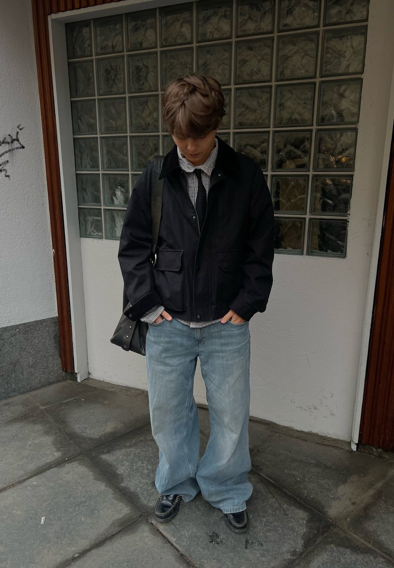 Person iført løstsiddende lyseblå jeans, sort jakke over ternet skjorte, sort slips, sorte sko, stående med hænderne i lommerne foran en væg af glasblokke.