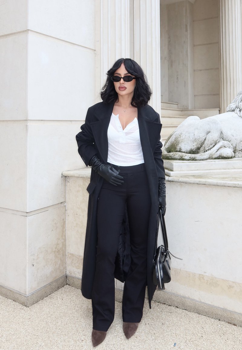 Femme en long manteau noir, gants noirs, pantalon noir, chaussures marron pointues et lunettes de soleil, debout près d'une statue de lion en pierre près d'un bâtiment à colonnes.