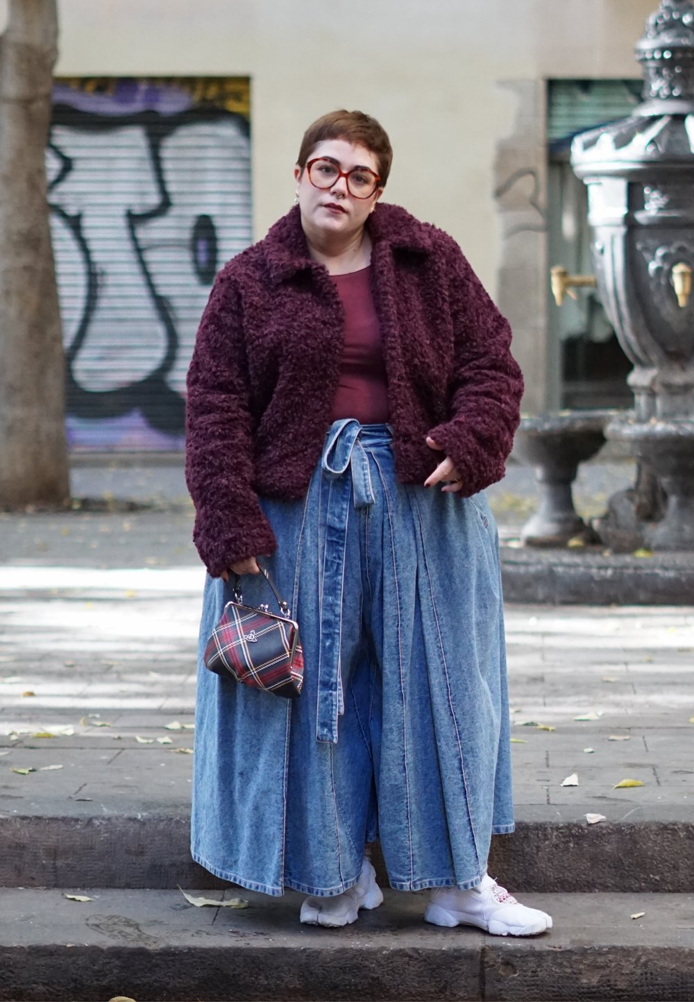 Personne portant une veste violette en tissu pelucheux, un pantalon large en jean bleu, des baskets blanches, tenant un petit sac à main à carreaux sur un trottoir en ville.