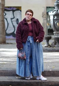 Personne portant une veste violette en tissu pelucheux, un pantalon large en jean bleu, des baskets blanches, tenant un petit sac à main à carreaux sur un trottoir en ville.