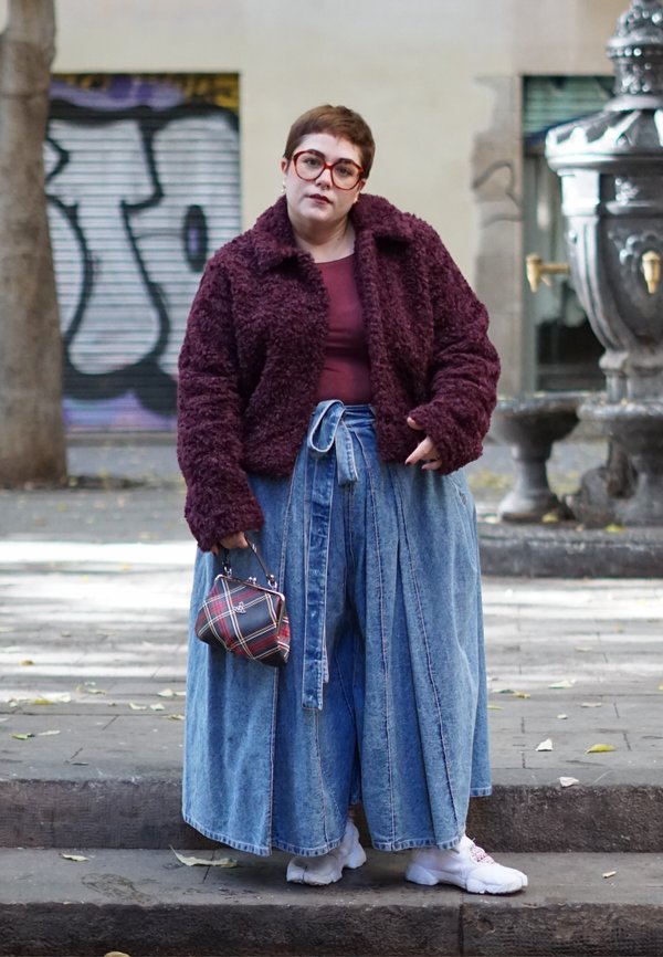 Osoba ubrana w fioletową, puszystą kurtkę, szerokie niebieskie dżinsy, białe trampki, trzymająca małą kratowaną torebkę na miejskim chodniku.