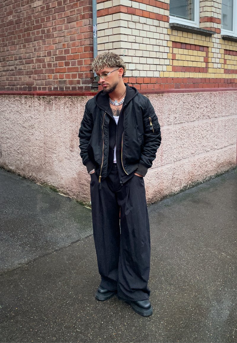 Giacca bomber nera con dettagli dorati, pantaloni neri oversize e stivali neri robusti. Maglietta bianca sotto e collana a catena d'argento. Capelli ricci.