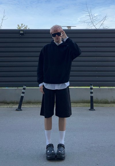Joven con cabello rosa se ajusta unas gafas de sol negras, viste sudadera negra con capucha, camiseta blanca, pantalones cortos negros, calcetines blancos y zuecos negros, de pie al aire libre.