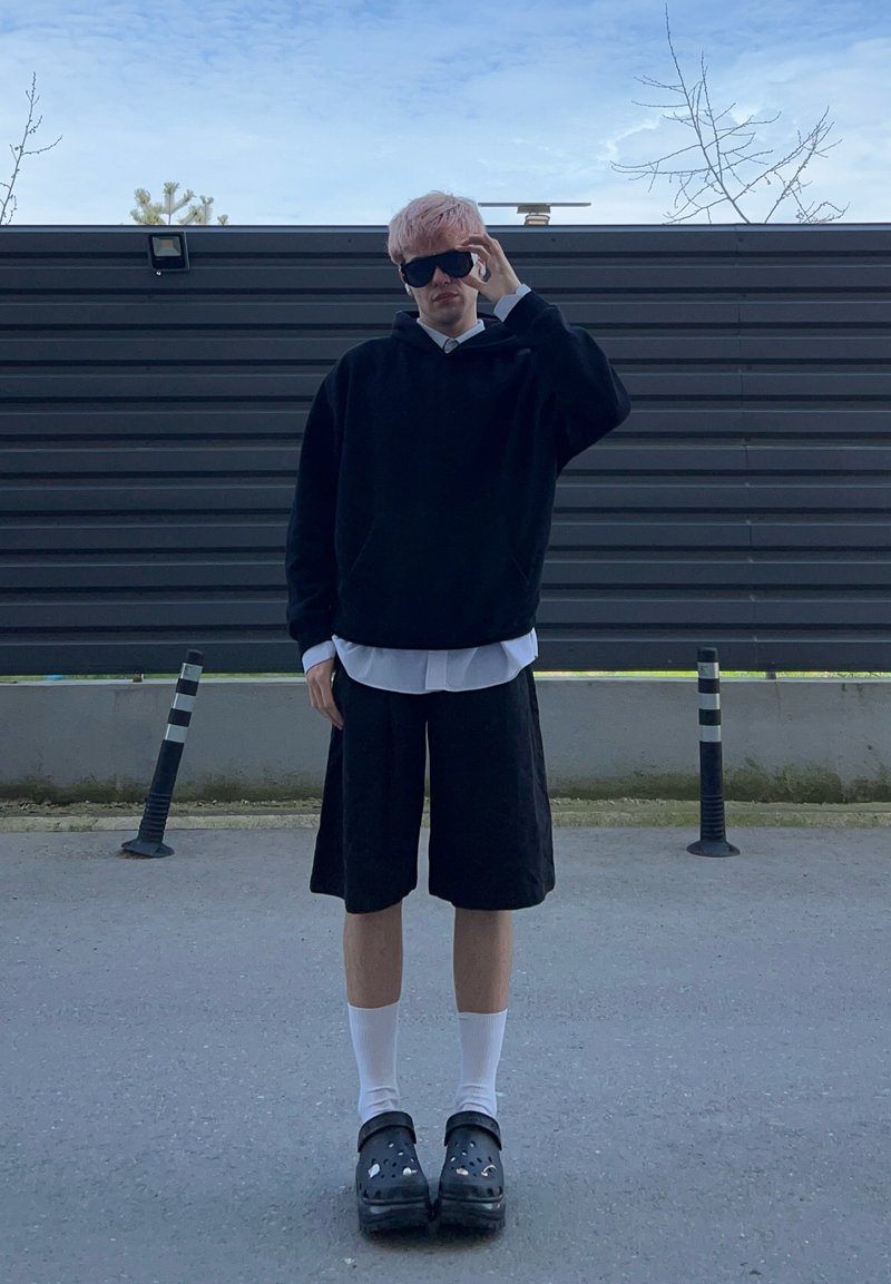 Jeune personne aux cheveux roses ajuste des lunettes de soleil noires, portant un sweat à capuche noir, une chemise blanche, un short noir, des chaussettes blanches et des sabots noirs, debout à l'extérieur.