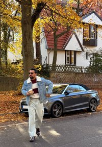 Mann i denimjakke og solbriller som går på en løvdekket gate med takeaway-kaffe i hånden, grå kabrioletparkert bil, høsttrær og hus i bakgrunnen.