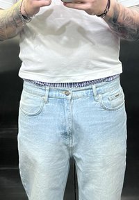 Jeans in denim azzurro chiaro con una finitura sbiadita, dotati di cinque tasche e chiusura a bottone argentato. Indossati con una maglietta bianca e una cintura a strisce.