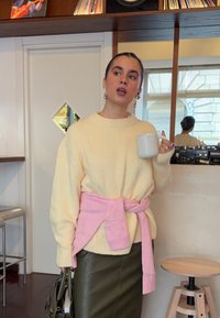 Overdimensjonert kremfarget genser med rosa belte i midjen, kombinert med en mørkegrønn skinnskjørt. Holder en hvit kopp, med store rundformede øredobber.