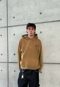 Sweatshirt capuz castanho com texto à frente, corte descontraído, combinado com jeans escuros que apresentam um porta-chaves na argola do cinto contra uma parede de betão texturizada.