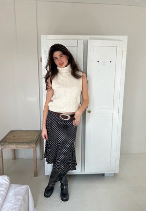 Suéter sin mangas de punto blanco, falda negra con lunares, cinturón marrón con hebilla de plata y botas negras hasta la rodilla. Fondo neutro.