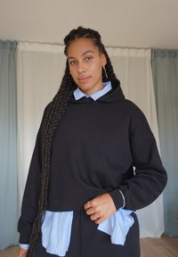 Sudadera con capucha negra de corte holgado, superpuesta a una camisa de cuello a rayas azul claro. Se ven trenzas y pendientes de aro dorados.