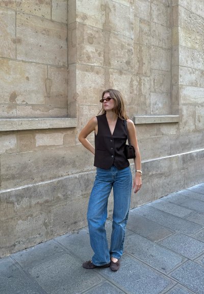 Chaleco negro sin mangas con botones, combinado con jeans de campana de color azul claro. Lleva un bolso pequeño oscuro y gafas, con un muro de piedra de fondo.