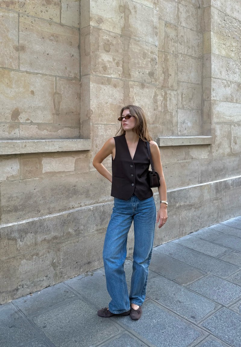 Gilet sans manches noir avec des boutons, associé à un jean flare bleu clair. Porté avec un petit sac noir et des lunettes, le tout devant un mur en pierre.